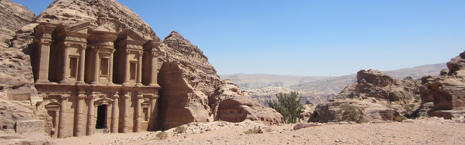 Ruins of a stone building with pillars against a rocky mountain. More rocky mountains are visible in the distance. 